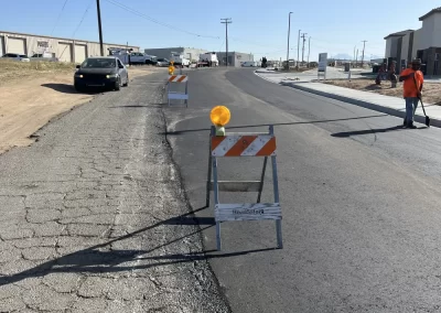 A newly paved road is blocked by barriers, with workers and vehicles in the background under a clear sky.