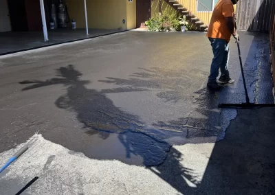 A man spreads fresh asphalt on a driveway outside a yellow apartment building on a sunny day.