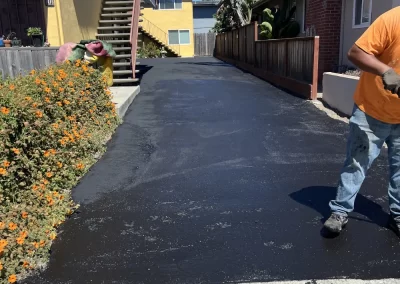 A freshly paved black asphalt driveway between two houses, with a worker standing on the right.