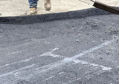 Worker in jeans and boots spreading fresh asphalt on a road with a large rake near a sidewalk.
