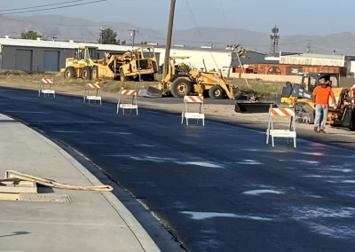 Freshly paved road with construction barricades; workers and equipment are visible in the background.