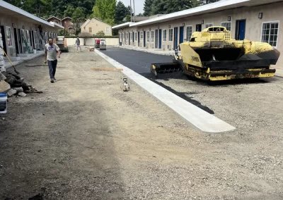 A person walks near freshly paved asphalt beside a yellow paving machine between motel buildings.