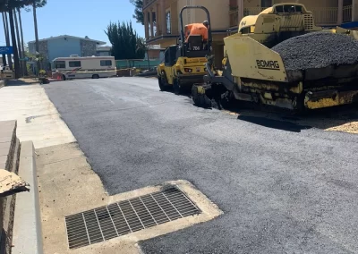 Construction vehicles paving a new asphalt road beside a building on a sunny day.