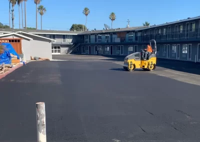 A worker drives a steamroller over freshly paved asphalt in a motel parking lot under a clear sky.
