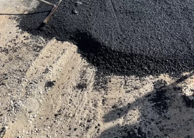 A rake spreads fresh asphalt over a gravel surface during road construction on a sunny day.