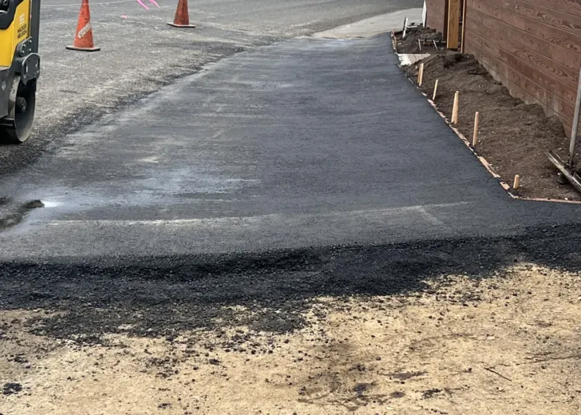 Freshly paved asphalt sidewalk with a raised edge meets a dirt path, orange cones in the background.