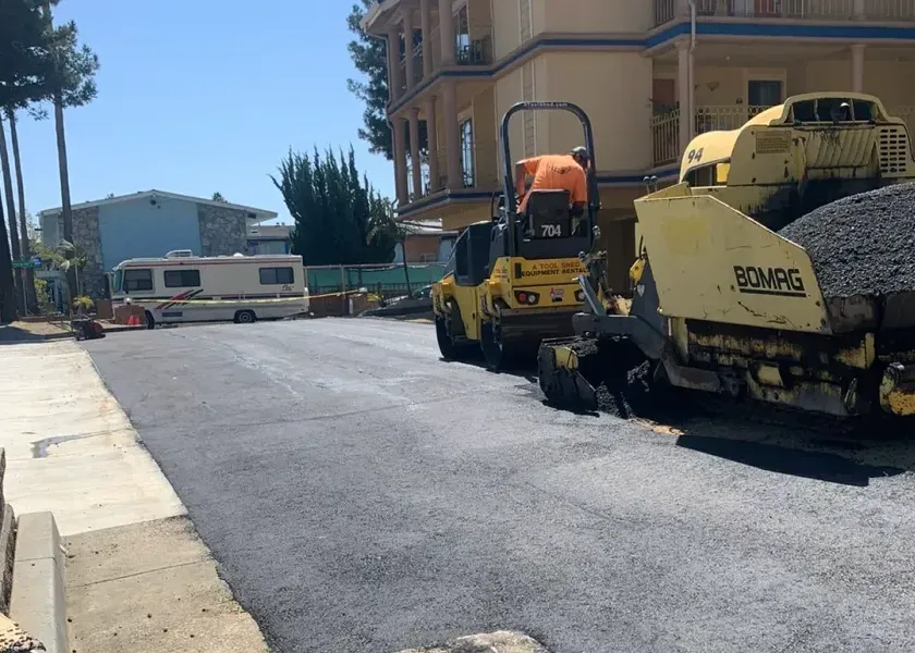 Paving machines laying fresh asphalt on a residential street near houses and an RV in the background.