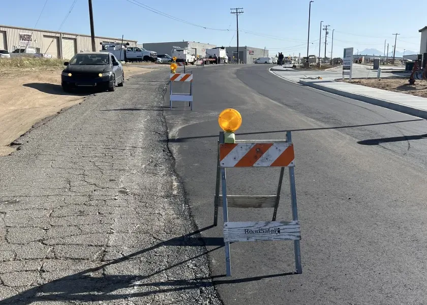 Road under construction with barricades blocking access; car stopped on the rough shoulder to the left.